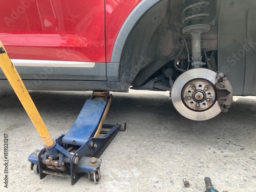 Low-angle view of a red car lifted by a hydraulic jack on a concrete floor. The wheel is removed, showing the brake disc and caliper assembly during a DIY repair or maintenance process.
