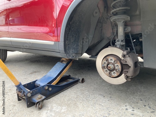 Low-angle view of a red car lifted by a hydraulic jack on a concrete floor. The wheel is removed, showing the brake disc and caliper assembly during a DIY repair or maintenance process.