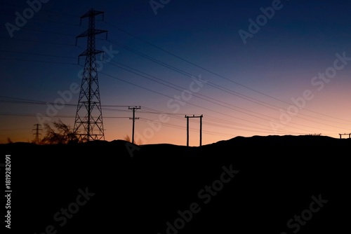 High Country Salmon area, New Zealand - May 23, 2025: A dramatic silhouette of power lines and transmission towers against a mountain backdrop during twilight. The sky transitions from orange to blue 