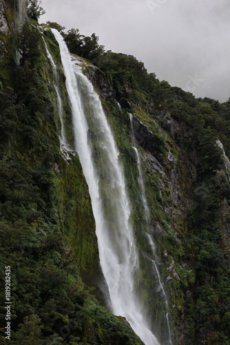 Milford Sound, New Zealand - May 20, 2025: A massive waterfall cascades down the sheer, moss-covered cliff face of the fjord. The scene is shrouded in mist and fog, typical of the Fiordland landscape.