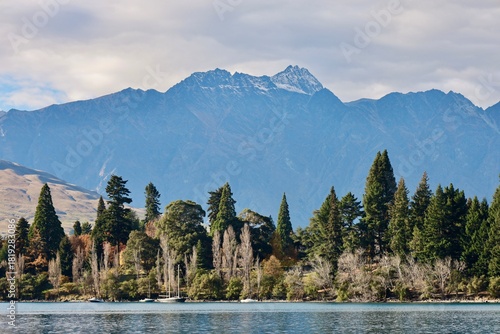 Queenstown, New Zealand - May 21, 2025: The snow-capped peaks of The Remarkables mountain range rise over Lake Wakatipu. The forested shoreline shows early autumn colors under a cloudy sky.