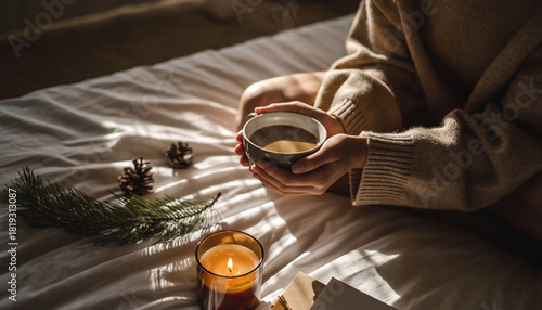 Manos con suéter beige sostienen una taza de café caliente sobre una cama blanca, en un ambiente cálido y acogedor con luces suaves y detalles naturales que evocan calma y confort.