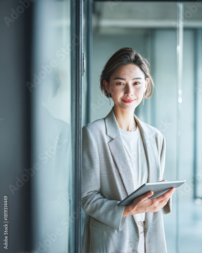 A confident woman in a stylish blazer holds a tablet, smiling in a modern office setting with glass walls. Free Download A confident woman in a stylish blazer holds a tablet, smiling in a modern office setting with glass walls. - GFXLoad