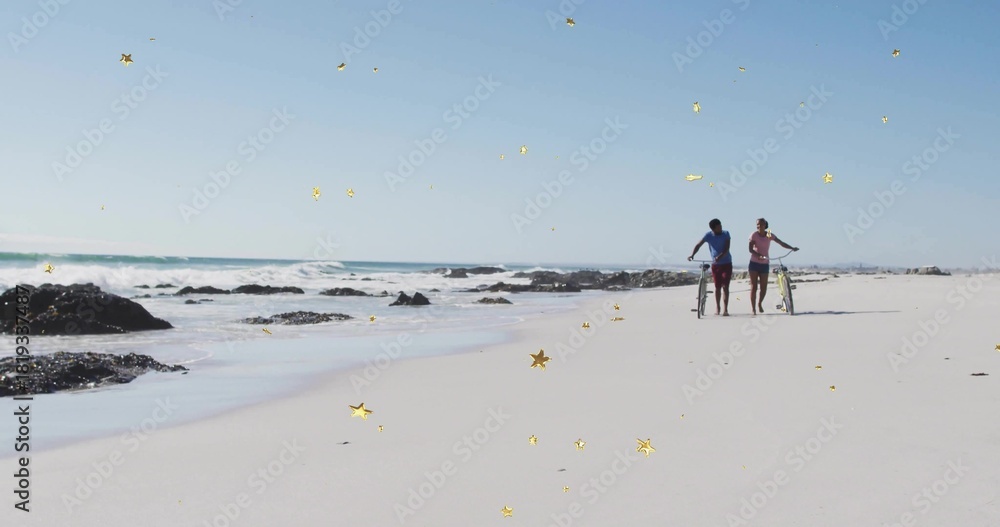 Naklejka premium Walking two adults pushing bicycles along sandy beach near surf and rocks, gold stars, copy space