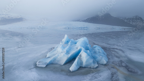 Aerial view of a striking blue iceberg adrift in the glacial lagoon, a serene dance of ice and water under a misty sky, Jokulsarlon, Sveitarfelagid Hornafjordur, Iceland.
