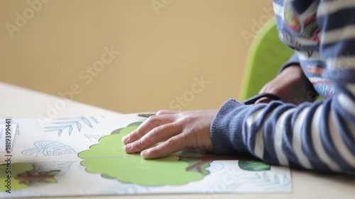 Close-up shot of a child's hands cutting a green tree image from paper using scissors. The kid is focused on the craft activity at a wooden table.

