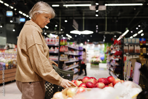 A woman selects apples from a display in a supermarket. Shelves filled with various products provide a vibrant backdrop. The lighting creates a warm atmosphere