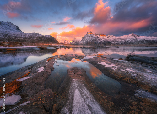 Arctic landscape with snowy mountains reflecting on calm lake with stones and colorful sky with clouds at sunrise. Winter in Lofoten islands, Norway. Sea, rocks in snow, pink sky with clouds at dusk