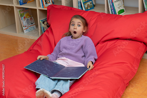 Young girl with surprised expression, sitting on a large red bean bag, reading an illustrated book in a cozy indoor library setting, showcasing joy and imagination
