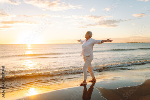 A joyful elderly woman feels free as she strolls along a sandy beach at sunset. Beautiful woman with gray hair enjoys stroll, walking barefoot along seashore. Concepts of nature, aging, retirement.