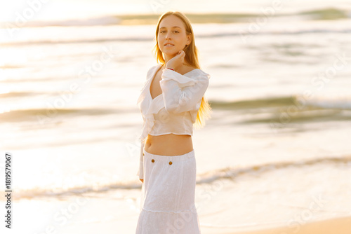 A happy woman in a white dress strolls along the beach at sunset. The young woman enjoys the seascape and feels the freedom of the outdoors. Concept of nature, enjoyment.