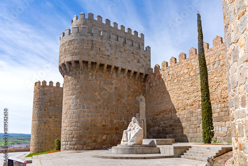 Foto A dramatic view of the Walls of Ávila with a white stone statue of Saint Teresa in the foreground, seated at the base of a medieval tower beside a tall cypress tree
