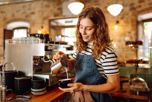 A female barista stands behind the counter with freshly brewed coffee in a cozy cafe. A smiling woman prepares and serves hot drinks inside. The concept is cozy, drinks, and small business.