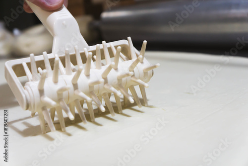 Close-up of a professional dough docker perforating pizza dough to prevent air bubbles during baking. Stainless-steel kitchen tool used in pizzerias and commercial kitchens. Close up. 