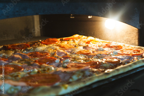 Large rectangular artisan pizza placed on a peel inside a professional electric oven. Freshly baked crust with visible texture, captured in a warm kitchen environment. Concept of bakery product