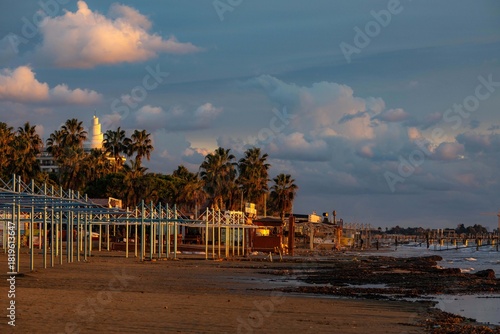 Fototapeta Naklejka Na Ścianę i Meble -  A serene, empty beach at sunset in low season, with seaweed-covered shore, bare sunbed frames, and palm trees silhouetted against the evening light. Side, Antalya, Turkey, Mediterranean.

