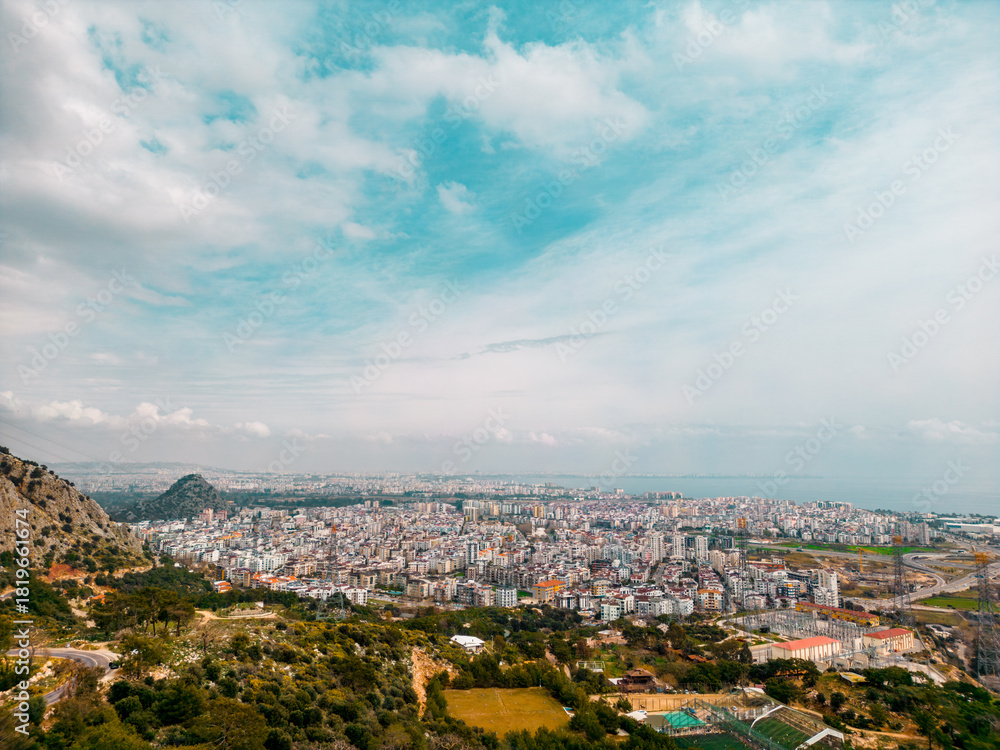 Fototapeta premium A serene aerial panorama of a large coastal city framed by soft blue skies, distant sea, and surrounding mountains. Natural daylight and calm atmospher