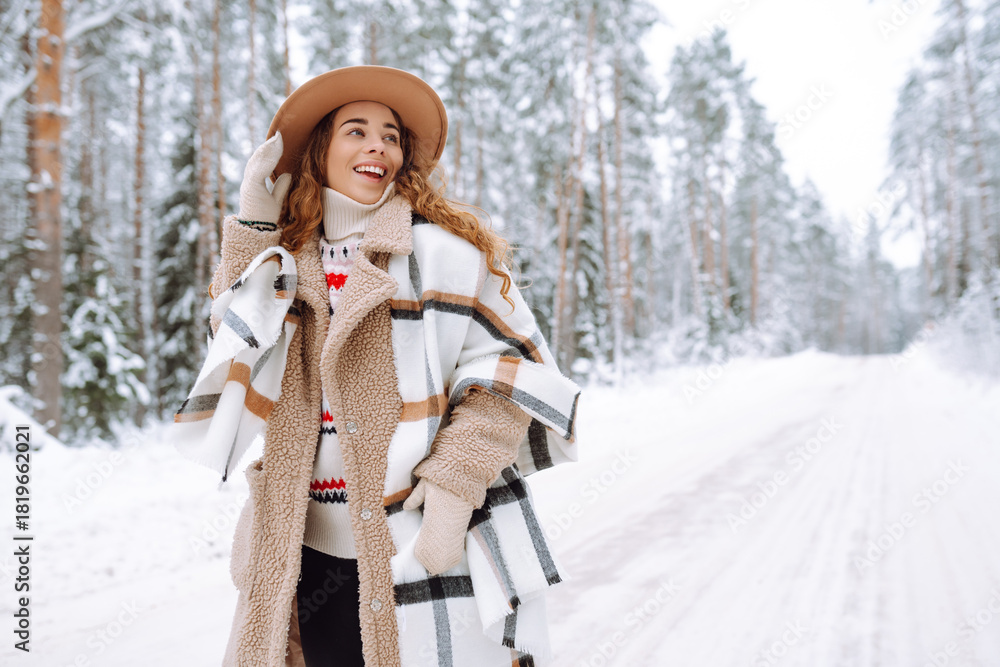 Fototapeta premium A cheerful woman stands on snowy path, wearing warm clothing. A hiker in a hat enjoys a snowy day outdoors in the forest. Nature, hiking, and lifestyle concepts.