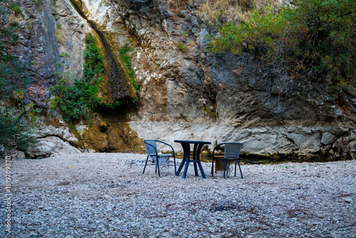 Empty chairs and table near small waterfall and rocky cliff surrounded by plants — quiet hidden spot in nature for relaxation and peaceful solitude