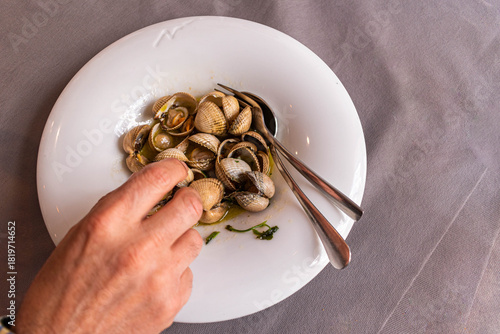 Hand reaching for fresh steamed cockles or clams with spoon on a white plate.
