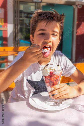 Boy eating strawberry ice cream in a large glass cup at an outdoor cafe on a sunny day.
