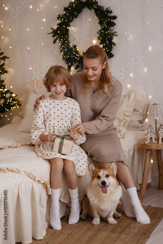 Wallpaper Mural Caucasian woman about 30 and girl about 7 in beige and white dresses sitting on bed opening gift, front view, corgi sitting below, cozy room with lights and wreath. Concepts: family, holiday. Torontodigital.ca