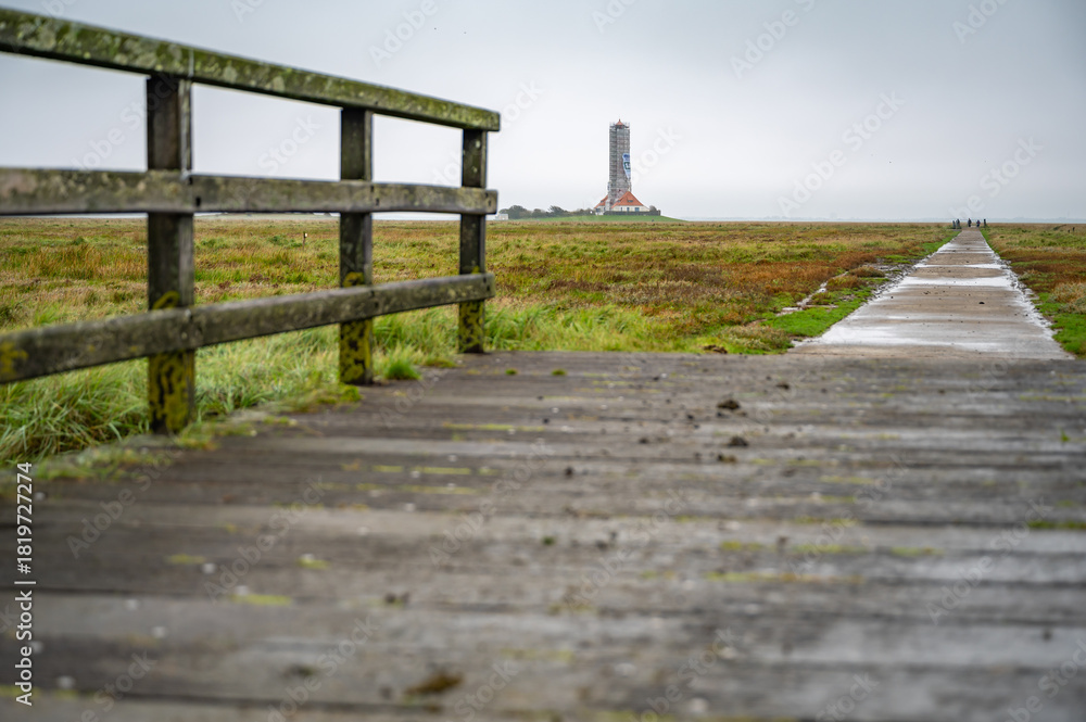 Obraz premium A weathered wooden walkway leads to Westerhever Lighthouse construction site in a grassy field, with a few people visible in the distance, North Sea, Germany