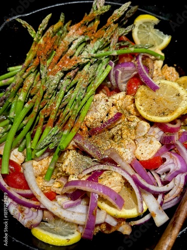 A cast iron pan full of colorful vegetables, lemons, and chicken, cooking together topped with spices. The vibrant colors of the red onions, tomatoes, asparagus, and lemons are very appetizing. 