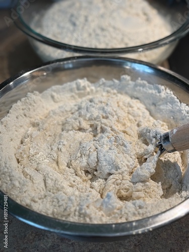Two mixing bowls full of the ingredients to make loaves of sourdough bread. The dough is in the process of being mixed and lots of lose flour is still visible in the bowls. 
