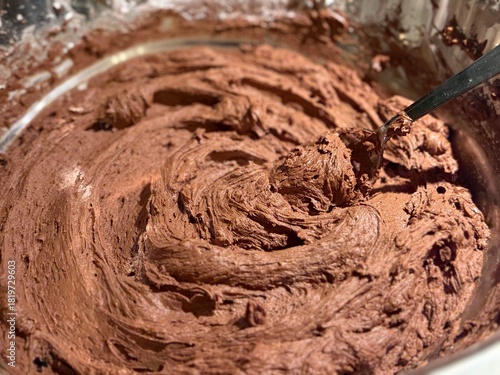 A stainless steel bowl full of smooth chocolate buttercream frosting. A spoon is in the middle, ready to scoop out the sugary sweet topping. 