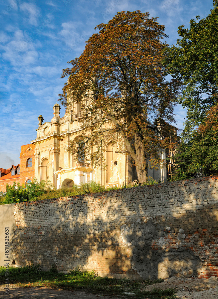 Fototapeta premium Historic Carmelite church in Bilshivtsi, Ukraine, with autumn foliage. The church sits atop a stone wall, bathed in golden sunlight against a blue sky with wispy clouds