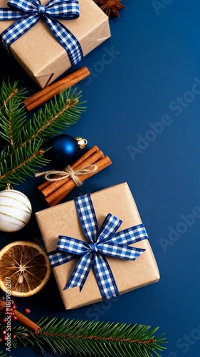 Top-down view of festive Christmas decorations with gift boxes, blue ornaments, cinnamon sticks and fir branches arranged on a blue background, creating a cozy holiday mood.