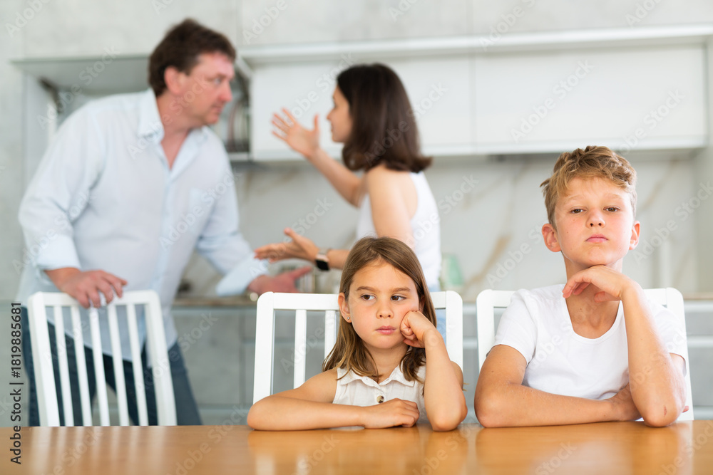 Fototapeta premium Children are sitting at kitchen table and hear their parents swearing, talking about divorce. Husband and wife are shouting at each other. Behind children are screaming parents in background, blurred