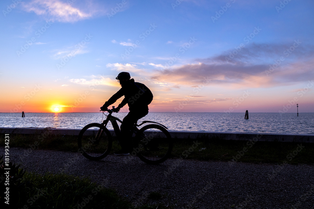 Obraz premium Silhouette of a cyclist riding along the waterfront at sunset on Pellestrina Island, Venetian Lagoon, Italy