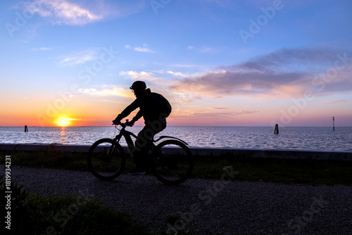 Silhouette of a cyclist riding along the waterfront at sunset on Pellestrina Island, Venetian Lagoon, Italy