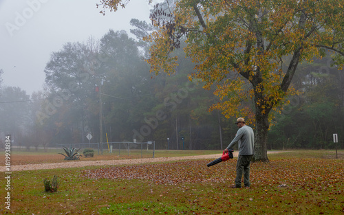 A person is seen blowing leaves in a yard on a foggy day. The autumn leaves are scattered on the ground, adding to the serene atmosphere.