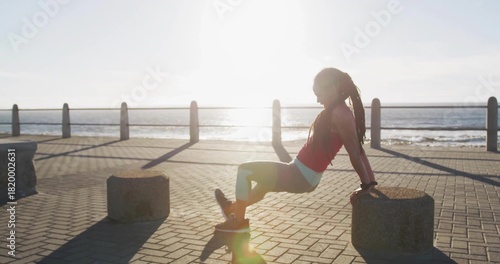 Doing triceps dips woman wearing red top, shorts, leggings on paved seaside promenade using bollard