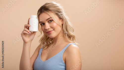 Young woman holding a white supplement bottle against a beige background while smiling, presenting a cheerful and confident wellness-focused pose suitable for health and lifestyle themes.