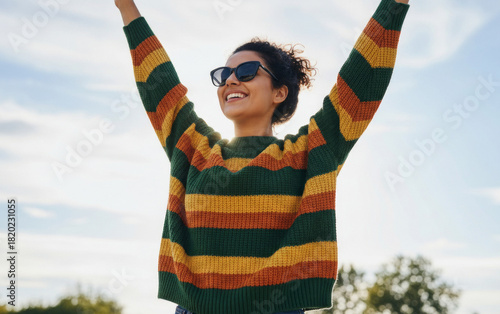 Woman in a striped sweater celebrates outdoors against a bright sky background, expressing joyful energy and freedom in a lively uplifting moment of happiness and confidence.