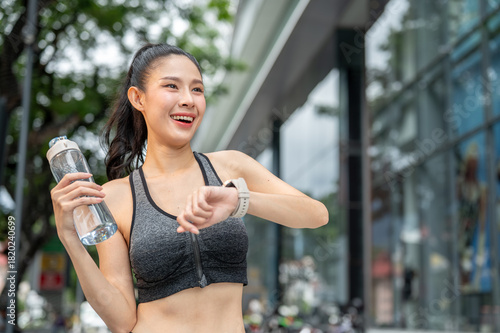 Asian woman in sportswear holding water bottle looking away from watch after exercising at mall park