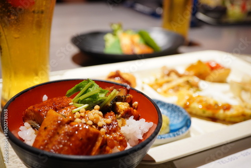 うなぎ丼とグラス、奥に料理が並ぶ店内での食事シーン。照りのあるうなぎと落ち着いた外食の雰囲気が伝わり、グルメ・旅行・レビュー用途に使いやすい写真。