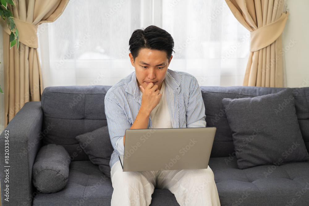 Obraz premium Man working on laptop at home sitting on sofa with thoughtful expression in bright living room