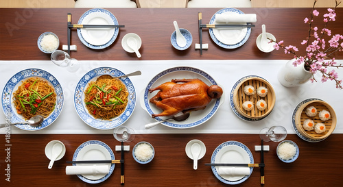 Overhead elegant Chinese banquet table with roasted duck noodles dim sum dumplings blue white tableware chopsticks and cherry blossoms festive family celebration Asian cuisine top view gourmet