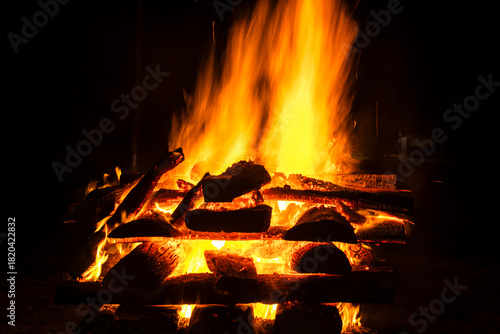 Narrow focus of a crackling bonfire at night in Brazil. Orange flames dance among stacked wood, illuminating the darkness. Sparks fly up, creating a fascinating visual spectacle.