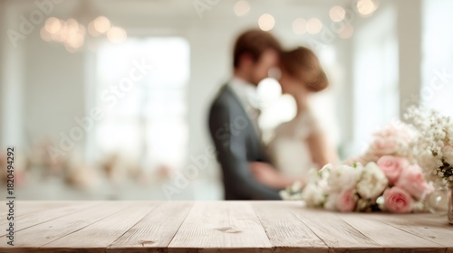 Wooden table foregrounds a romantic, blurred couple in wedding attire with elegant floral arrangements in a sunlit room.
