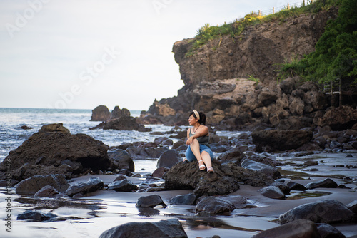 A contemplative woman sits peacefully on a large rock at the beach, gazing out at the ocean