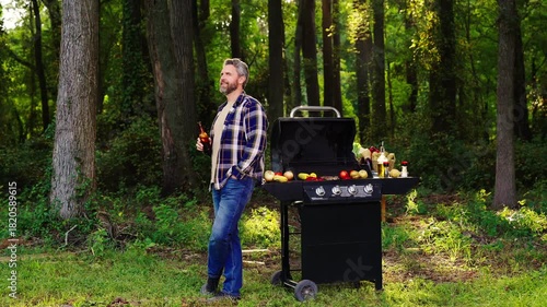 Middle-aged man cooking beef on the grill outdoors. Relaxing with beer and barbeque in nature. Man with grill and drink beer. Man grilled food in backyard. Man with bottle beer cooking near grill.