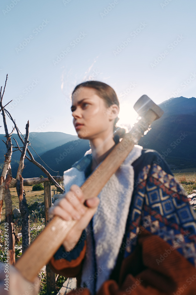 Fototapeta premium Woman hammer outdoors mountains portrait rural field sledgehammer, young laborer holding tool in backlit landscape, sunlight on patterned jacket, focused expression and workwear detail.