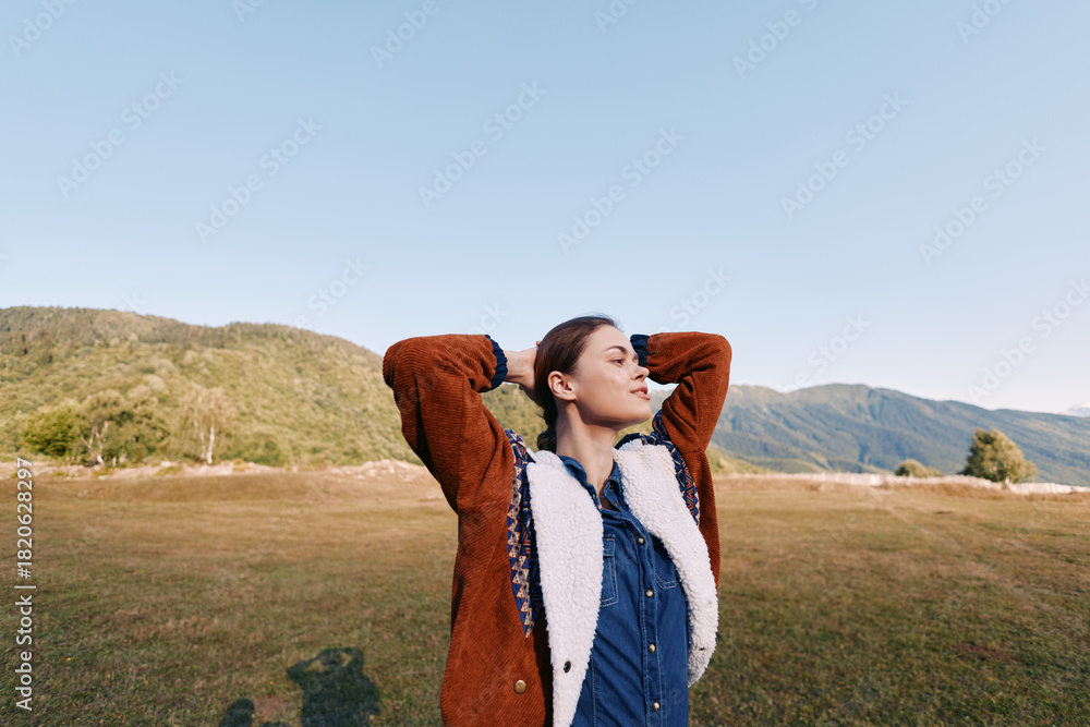 Fototapeta premium Woman in autumn jacket stands in meadow with mountains in background, portrait of relaxation and outdoor lifestyle, enjoying fresh air and scenic nature landscape.