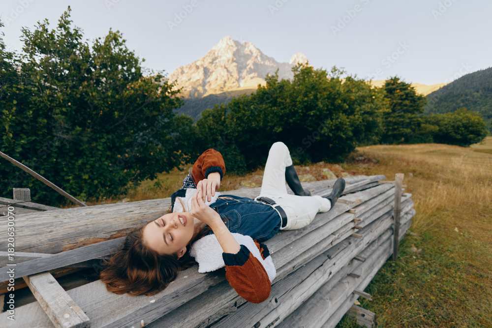 Fototapeta premium Woman on wooden fence using phone, relaxing in meadow with mountains in background and countryside nature. Casual sweater, rural leisure and peaceful outdoor lifestyle portrait.